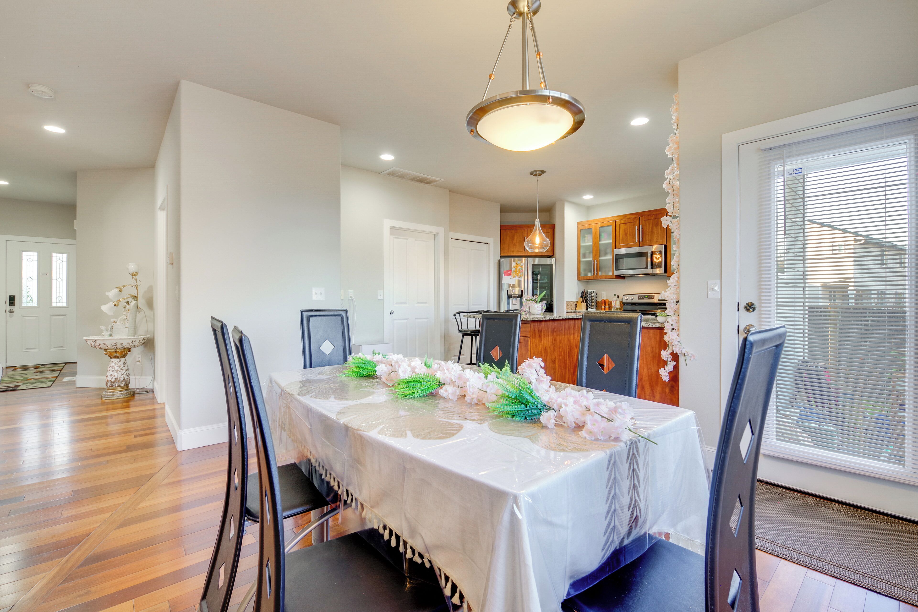 A dining area with a table set for four, surrounded by chairs. A kitchen is visible in the background with wooden cabinets and a microwave.