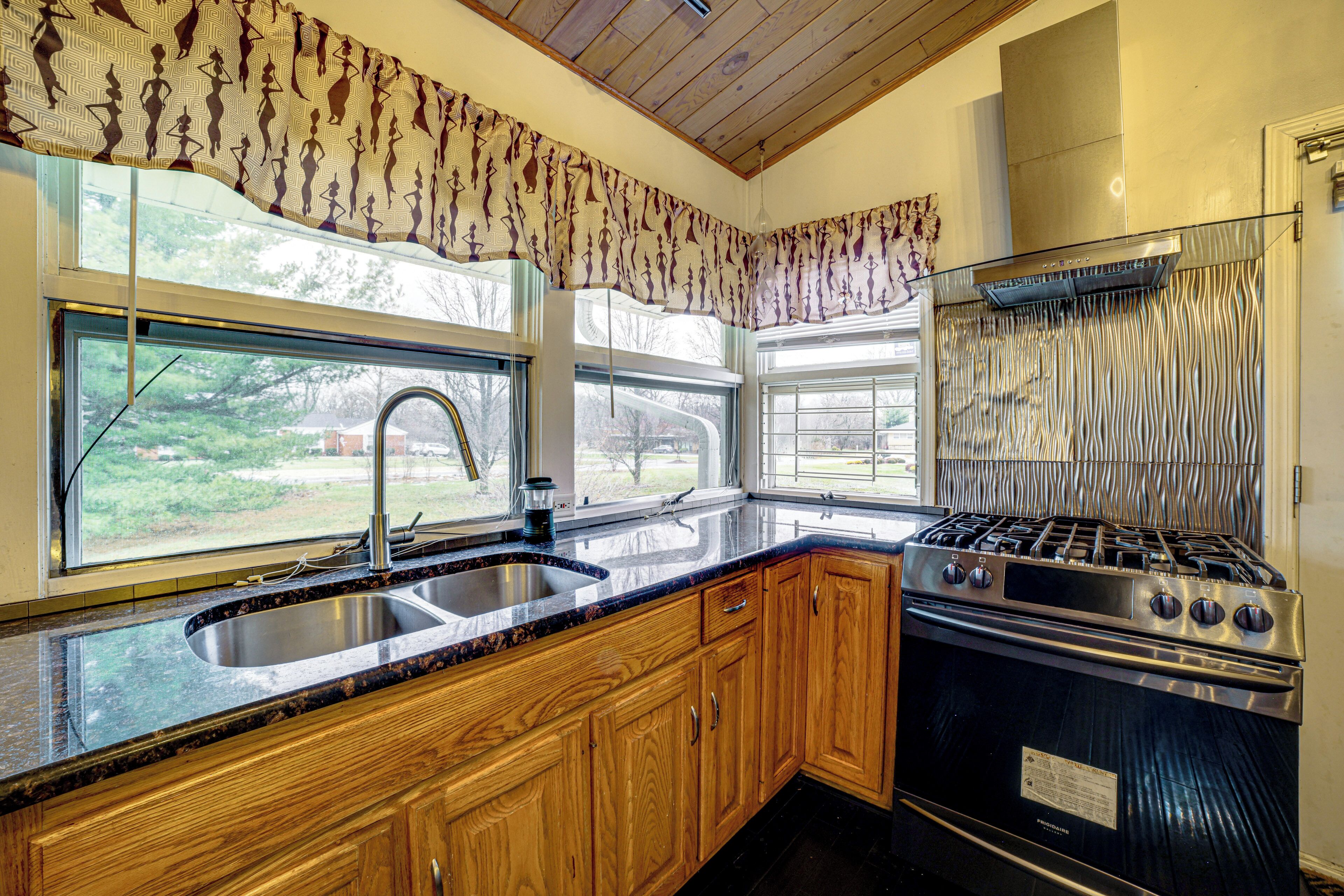 A kitchen with wooden cabinets, a stainless steel sink, a stove, and a window with a view of the outdoors.