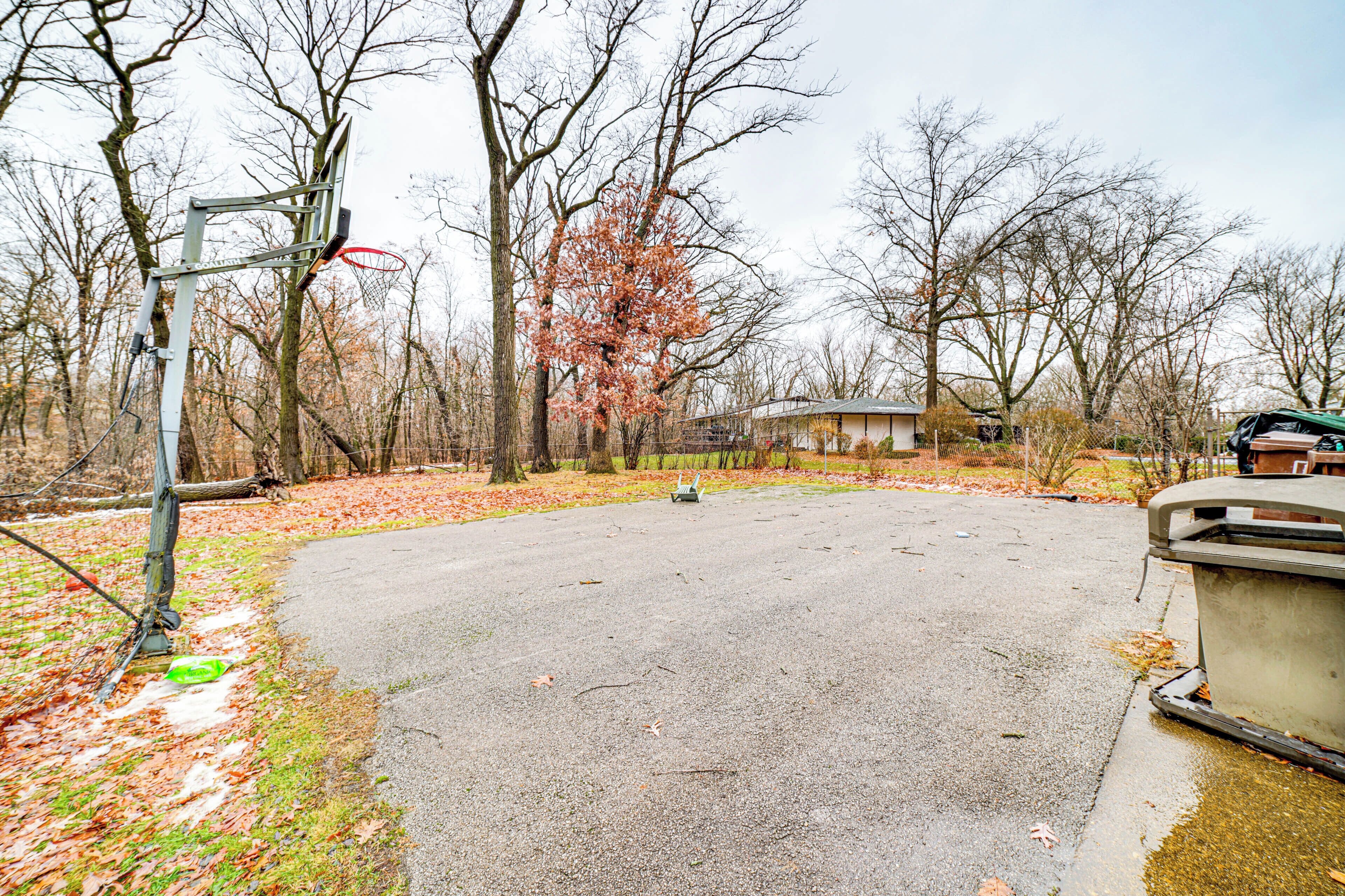 A parking area with a basketball hoop, surrounded by trees and a building in the background.