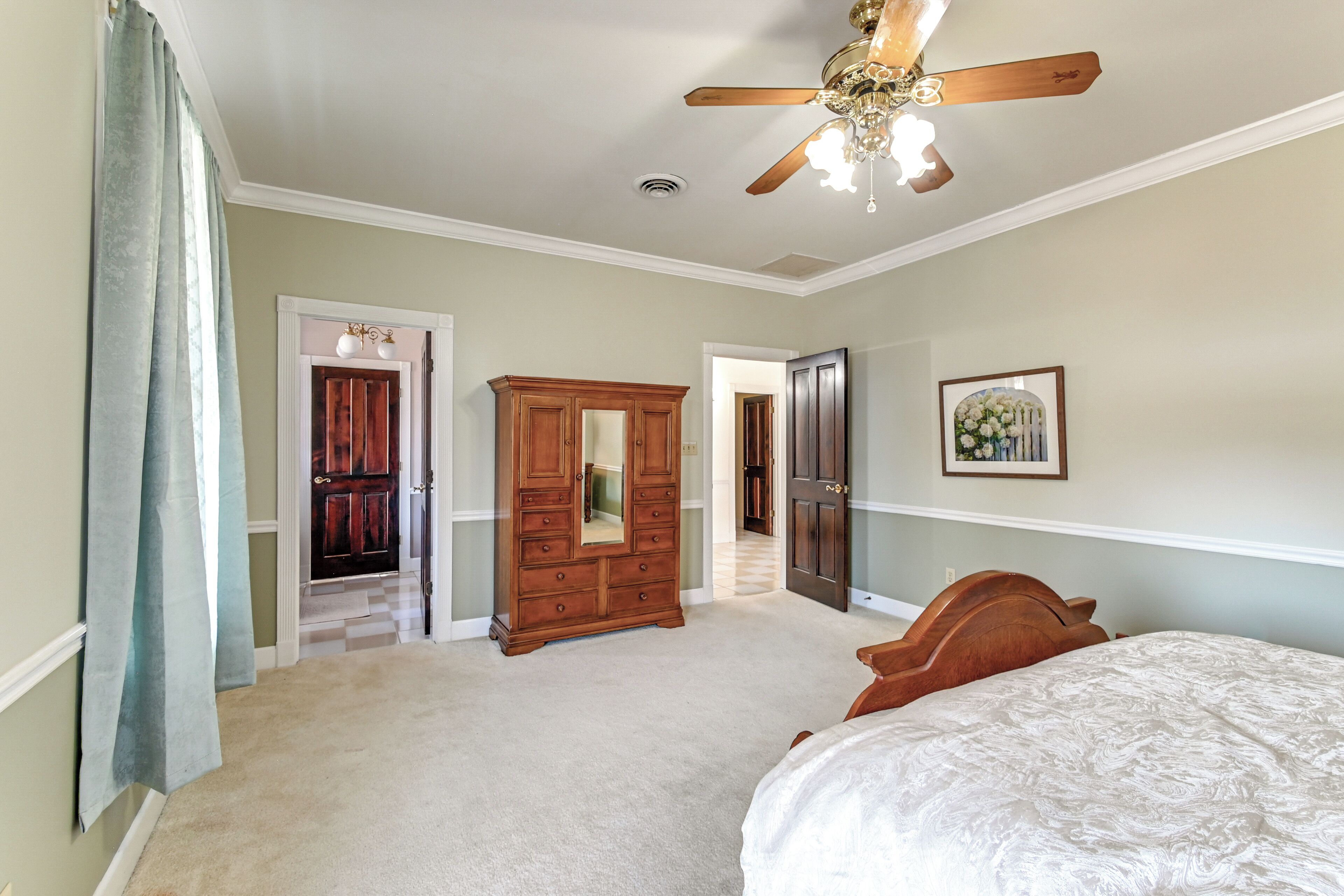 A bedroom with a bed, a wooden wardrobe, a ceiling fan, and a door leading to another room.