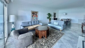 A modern living room with a grey sofa, a blue sofa, a wooden coffee table, and a patterned rug. There is a dining area with a table and chairs, and a large window providing natural light. - Cliff Apartment With sea View in Sidi Bou Saïd (Sidi Bou Said)