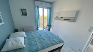 A bedroom with a bed, a window with curtains, a radiator, a wall-mounted picture, and a door leading to another room. - Charming 1-Bedroom with Balcony, Parking Included, Close to Beach & Shops (Arcachon)