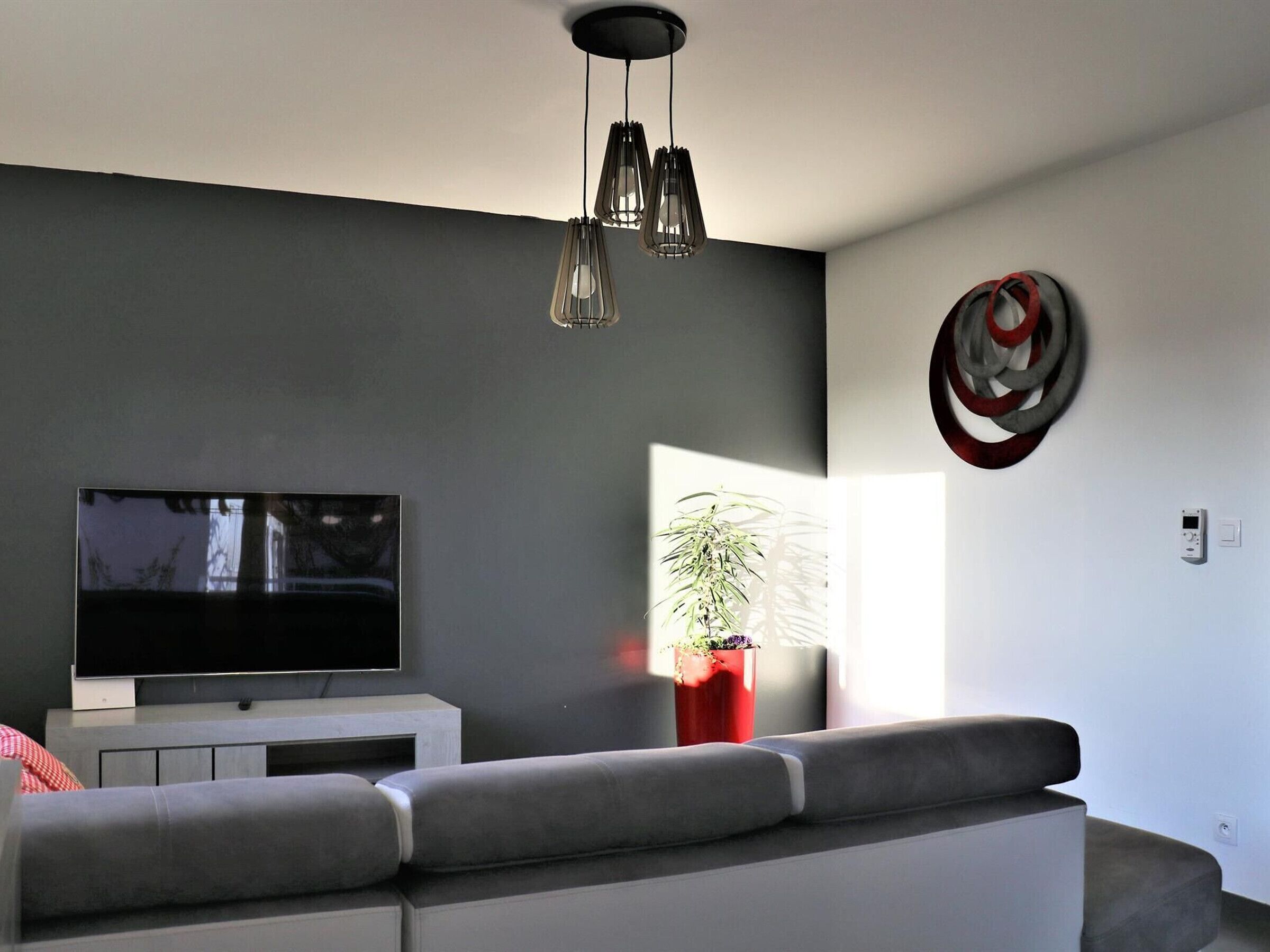 A modern living room with a flat-screen TV, grey sofa, pendant lighting, a red vase with a plant, and a circular wall decoration.