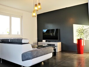 A modern living room with a large flat-screen TV, a grey sofa, a coffee table, and a red potted plant. - Spacious family home, pets allowed in Oberentzen (Oberentzen)