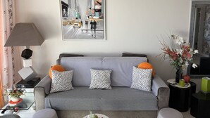 A modern living room with a grey sofa, patterned cushions, a painting of a city street, a lamp, and a vase with flowers. - Charming T2 in Saint-Palais-sur-Mer, near Nauzan beach, private box (Saint-Palais-sur-Mer)
