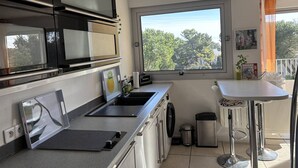 A modern kitchen with stainless steel appliances, a large window with a view of trees, and a small dining area with a table and chairs. - Charming T2 in Saint-Palais-sur-Mer, near Nauzan beach, private box (Saint-Palais-sur-Mer)