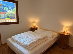 Un lit simple avec une literie blanche, deux tables de chevet avec des lampes, une fenêtre donnant sur un paysage de montagne et des bâtiments, et un plancher en bois.