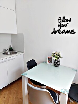 A modern kitchen with white cabinets, a stainless steel sink, and a glass-top table. A black chair is positioned at the table, and a decorative sign on the wall reads "follow your dreams." - Spin&Fly (Zagreb)