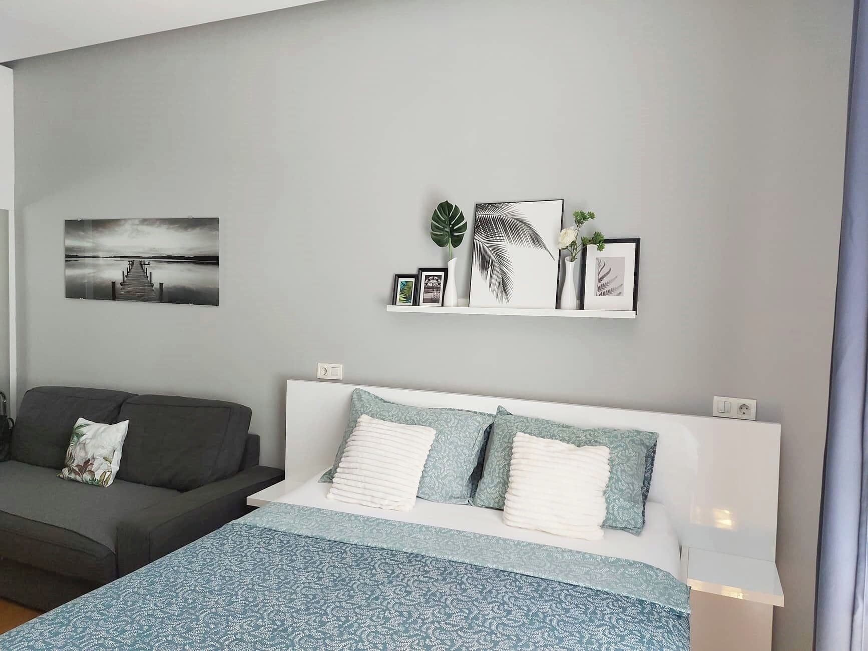 A modern bedroom with a bed, grey walls, a grey sofa, and a wall-mounted shelf with framed pictures and plants.