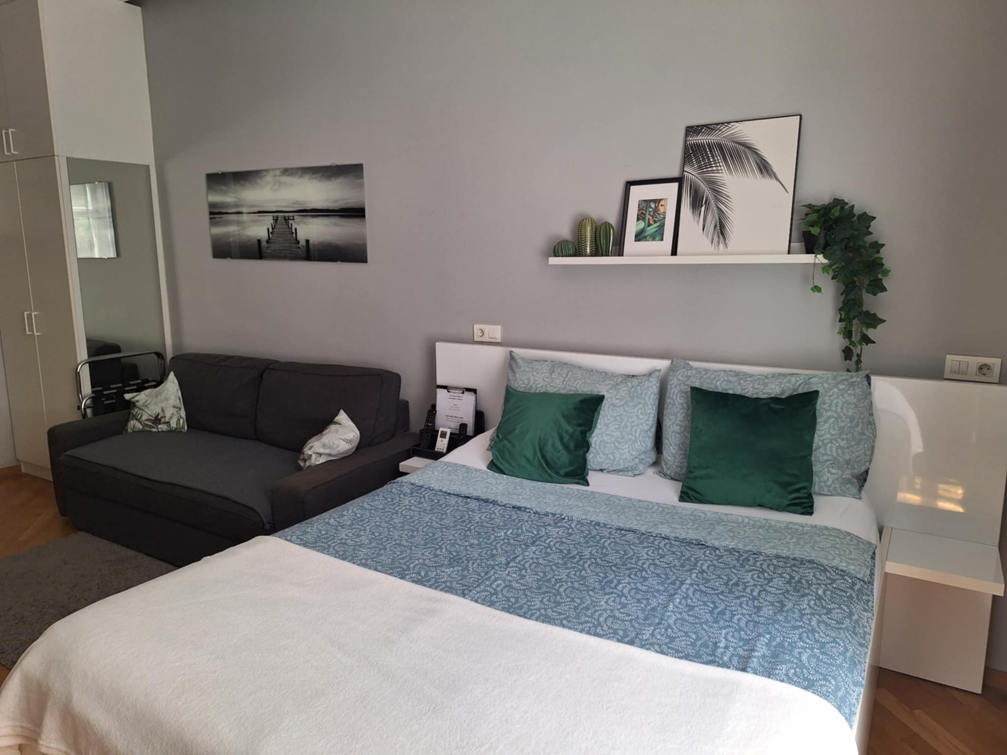 A modern bedroom with a bed, a sofa, a nightstand, and a wall-mounted shelf with decorative items.