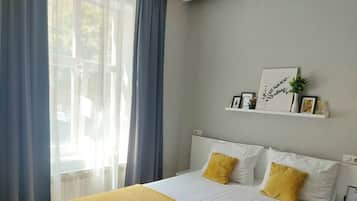 A neatly made bed with white and yellow bedding, a window with blue curtains, and a wall-mounted shelf with decorative items.