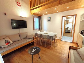 A cozy living room with a sofa, a dining table with chairs, a TV mounted on the wall, and a small round table with a sign reading "GRANDE PENTE 9 LA PISTE". - Charming 2-room apartment in Méribel, 4 people, near slopes (Méribel)