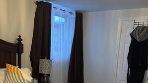 Ett sovrum med en stor säng, ett nattduksbord, en lampa, ett fönster med gardiner och en dörr med en klädhängare.