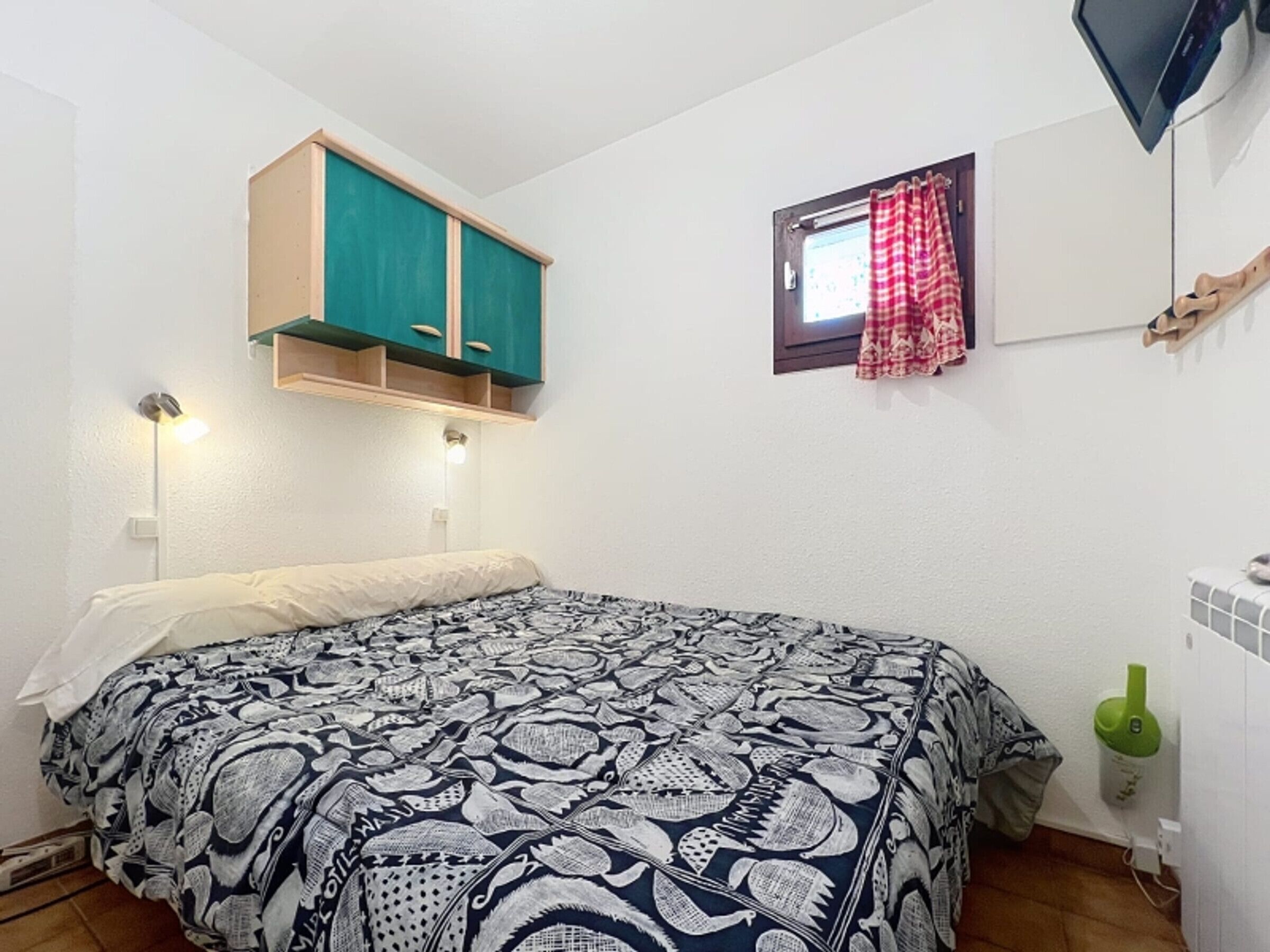 Een slaapkamer met een bed, een klein raam met rode gordijnen, een groen kastje boven het bed en een aan de muur bevestigde televisie.