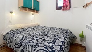 Een slaapkamer met een bed, een klein raam met rode gordijnen, een groen kastje boven het bed en een aan de muur bevestigde televisie.