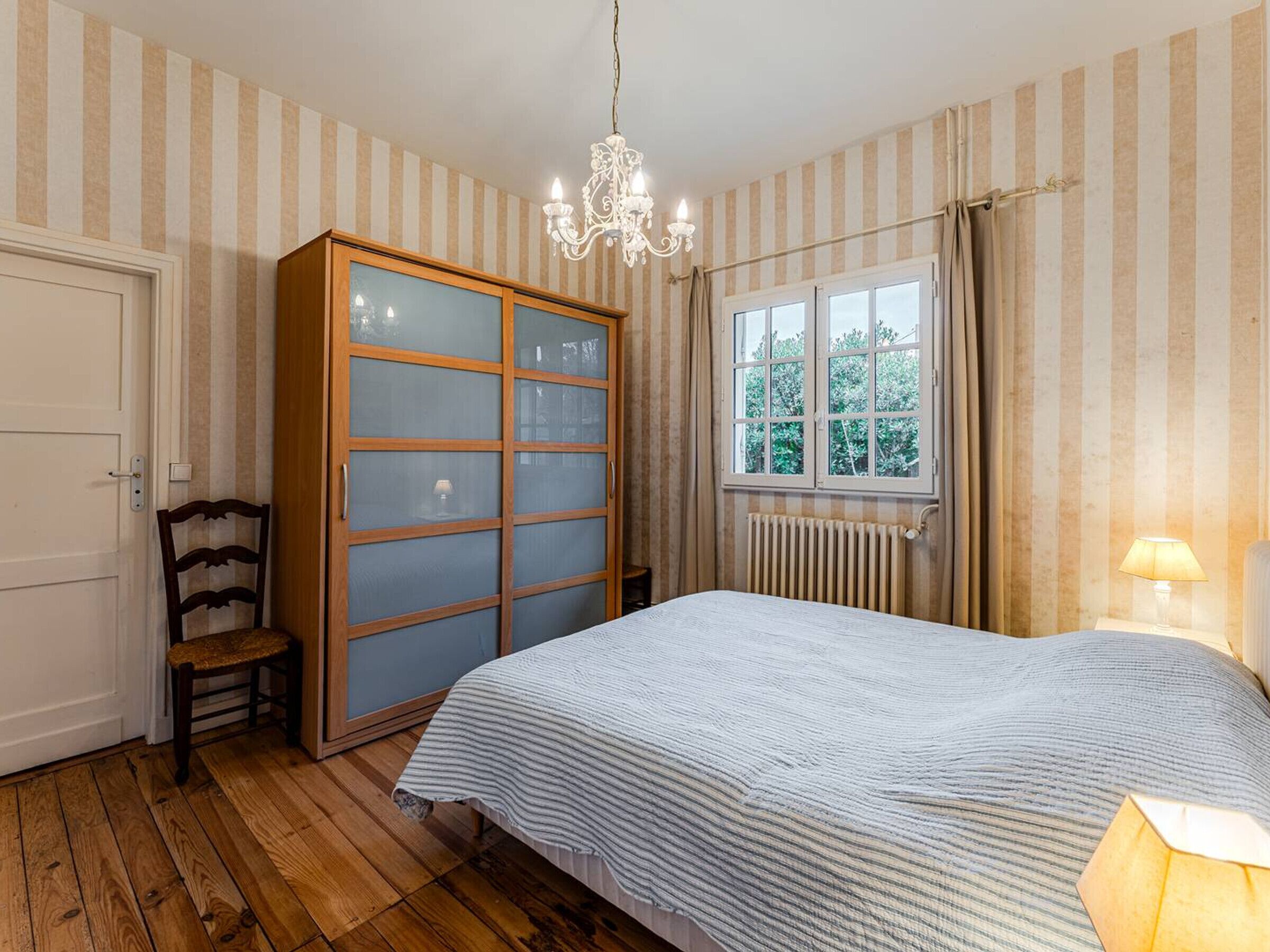 Ein Schlafzimmer mit Holzschrank, einem Bett mit gestreifter Bettdecke, einem Sessel, einem Fenster mit Vorhängen und einem Heizkörper.