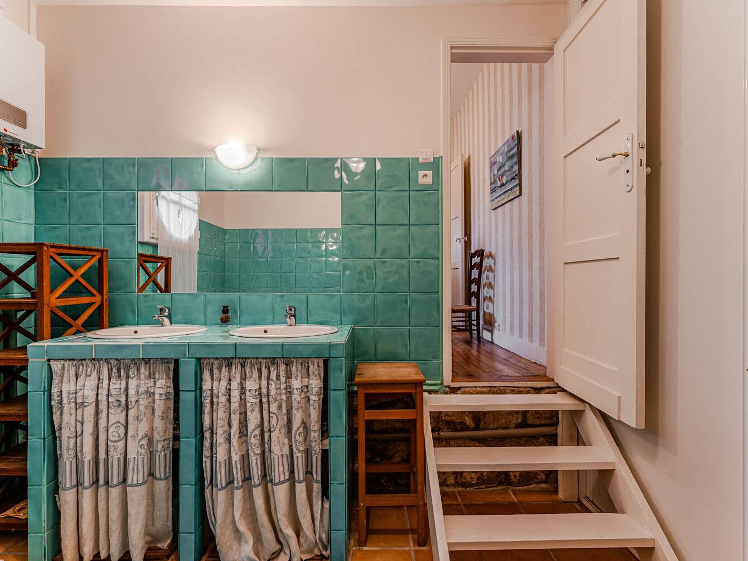 A bathroom with teal tiles, two sinks, wooden chairs, a mirror, and a staircase leading to another room.