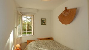 Ein Schlafzimmer mit einem Einzelbett, einem Nachttisch mit Lampe, einem Fenster mit Blick auf Grünflächen, einem Wandventilator und einem gerahmten Bild.