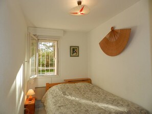 Un dormitorio con una cama individual, una mesita de noche con lámpara, una ventana con vistas a zonas verdes, un ventilador de pared y un cuadro enmarcado.