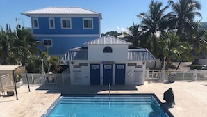 Uma piscina retangular com água azul cristalina, rodeada por um deck revestido de azulejos. Ao fundo, um edifício com fachada azul e branca, e palmeiras visíveis.