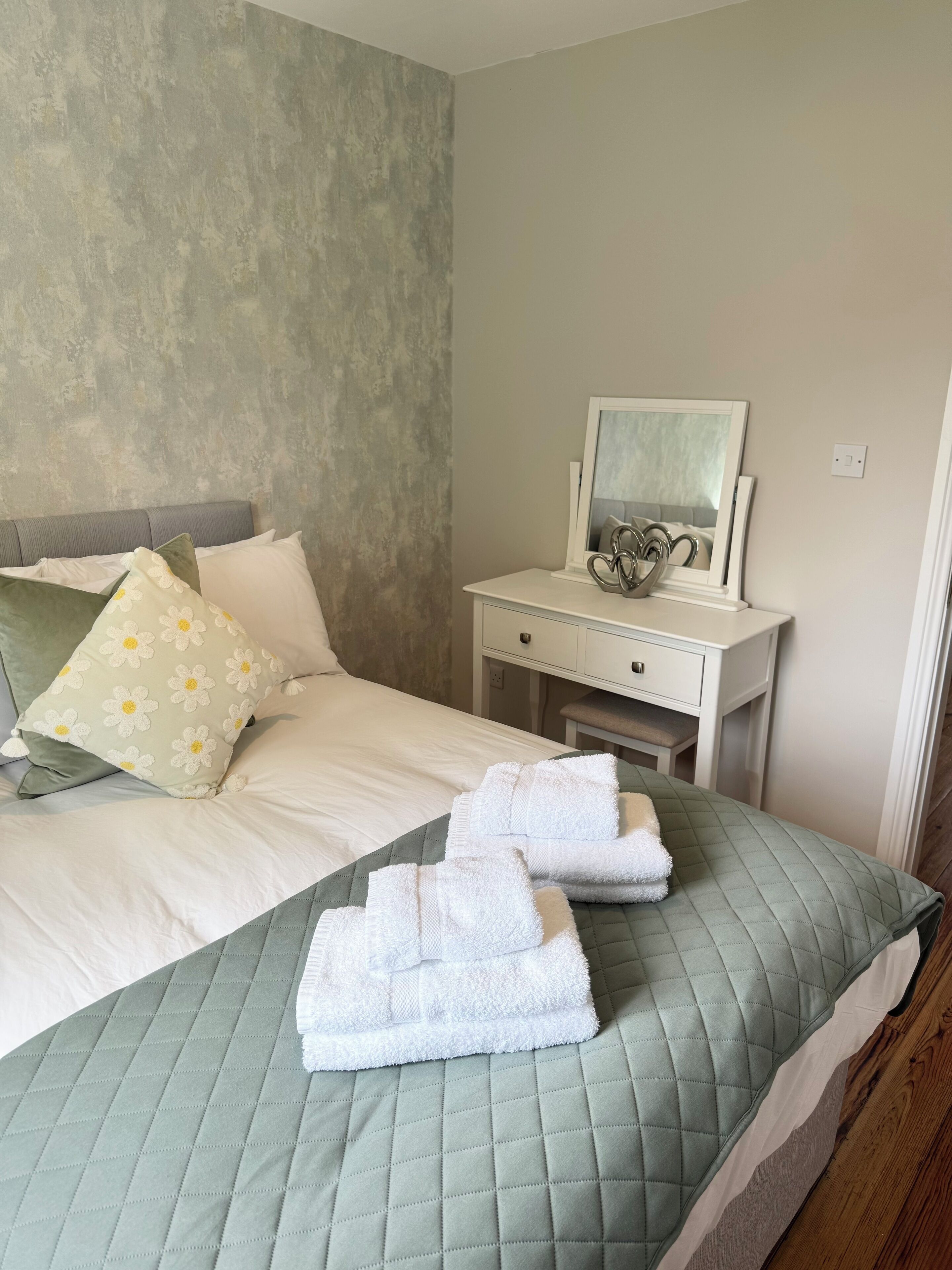 A neatly made bed with a quilted green bedspread and white pillows, a bedside table with a mirror and decorative items, and a textured grey wall.