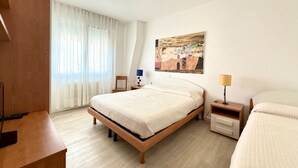 Moderni hotellihuone, jossa on puinen sänky, pieni yöpöytä, yölamppu, tuoli ja suuri ikkuna, jossa on ohuet verhot.