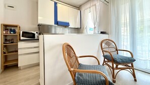 Kompakti keittiö, jossa on valkoiset kaapit, mikroaaltouuni ja rottinkituoli, jossa on pehmuste.