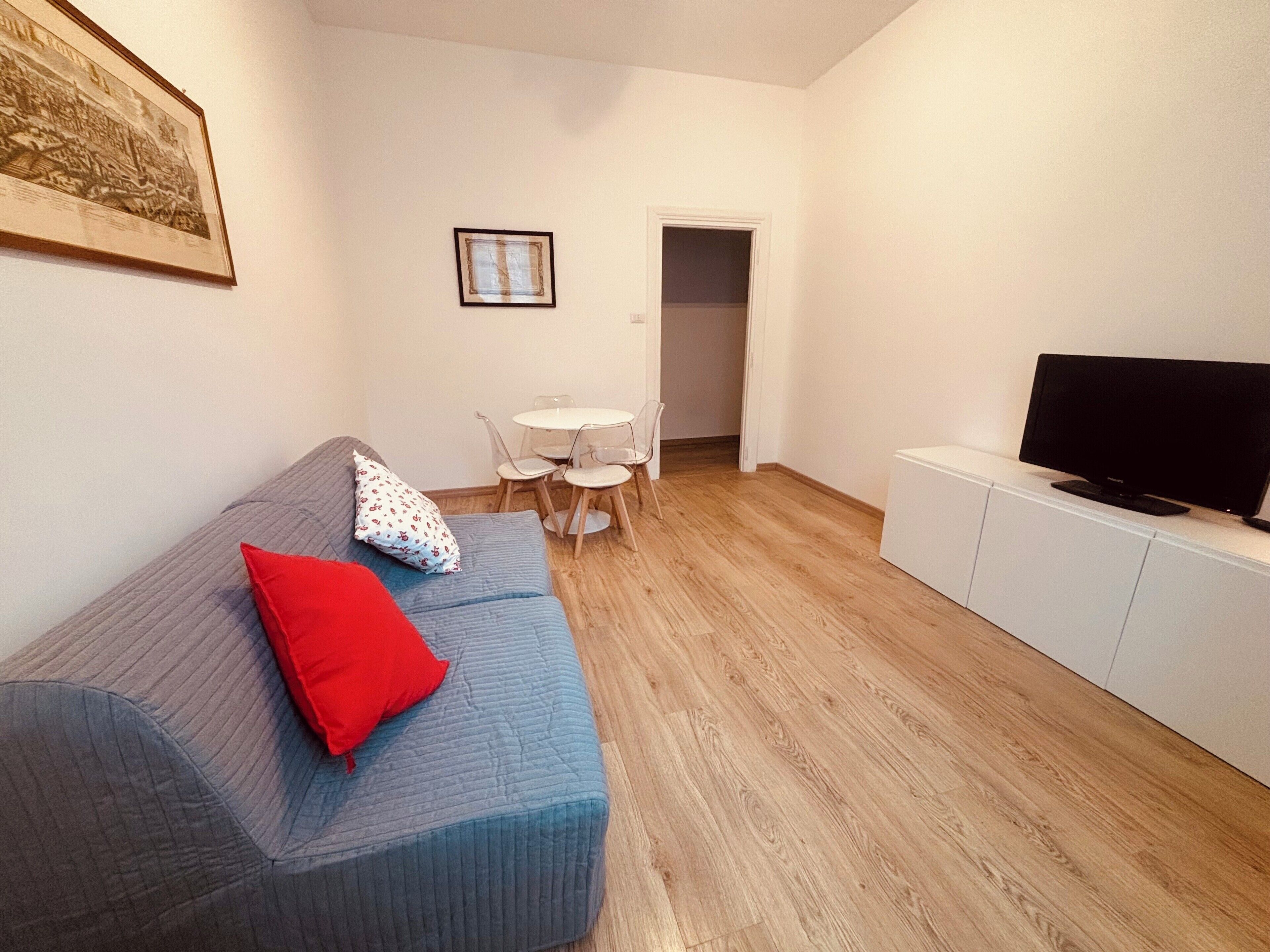 Un salon avec un canapé bleu, des coussins rouges et blancs, une petite table blanche avec des chaises, un téléviseur posé sur un meuble blanc et un tableau encadré accroché au mur.