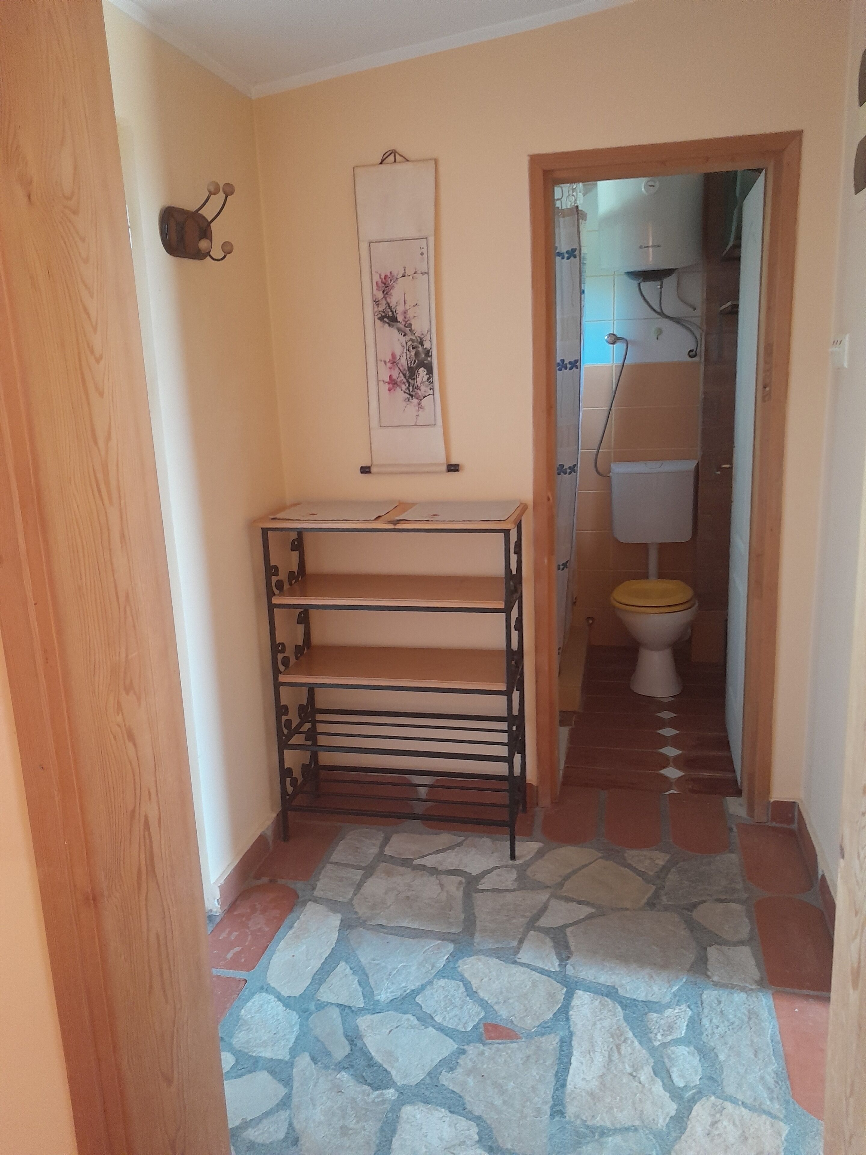 Een smalle gang met een betegelde vloer, een metalen rek met planken, een kunstwerk aan de muur, en een toilet zichtbaar door een geopende deur.