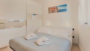 Une chambre d’hôtel avec un lit, un grand miroir, un tableau représentant un paysage côtier et un rideau blanc.