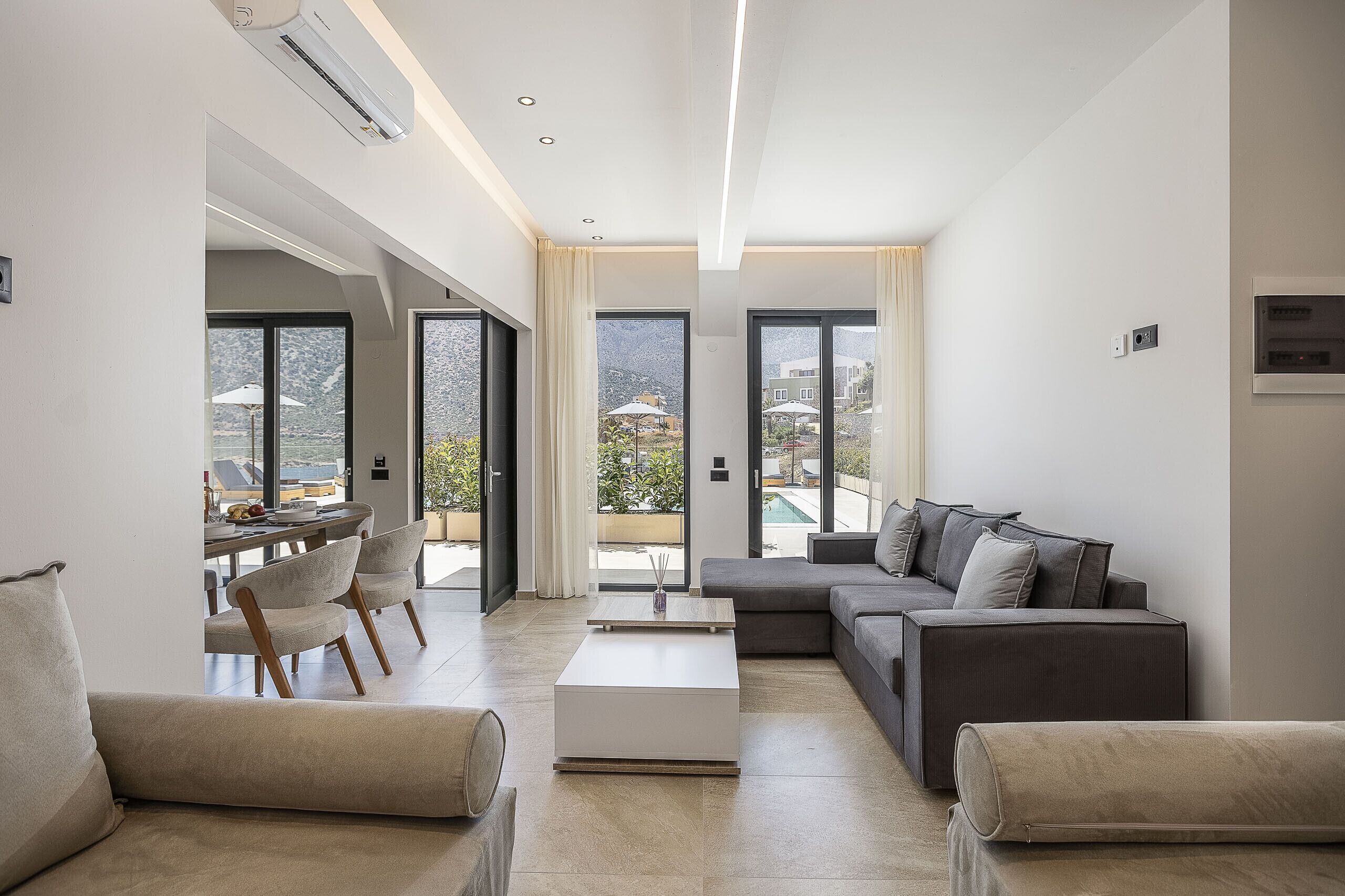 A modern living room with a sofa, coffee table, and chairs. There are large windows with curtains, an air conditioning unit, and a view of the outdoors.