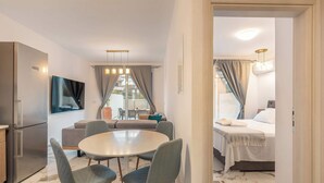 A modern hotel room with a bed, a dining area with a round table and chairs, a kitchen area with a refrigerator, and a large window with curtains. - Punta Mare Villas - Adults Only (Pigadia)