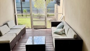 Un porxo cobert amb sòl de fusta, dos sofàs, una taula de centre i una porta corredissa de vidre que dona a una zona exterior tancada per una tanca.