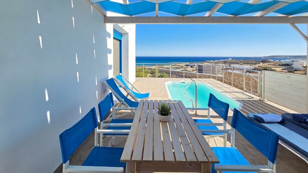 A covered outdoor area with a pool, blue lounge chairs, and a wooden table with a potted plant. - |    Rania Villas   | (Kipi Afiarti)