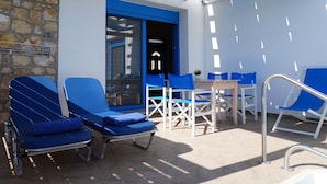 A sunlit outdoor seating area with blue lounge chairs and a table, stone wall, and a blue-framed door. - |    Rania Villas   | (Kipi Afiarti)