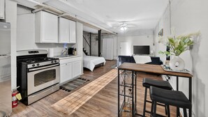 Une cuisine compacte avec des meubles blancs, une cuisinière et un évier. La pièce adjacente contient un lit recouvert d’une couette blanche.