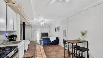 A modern kitchen with white cabinets, a stove, and a sink. There is a blue sofa and a small table with two stools in the living area.