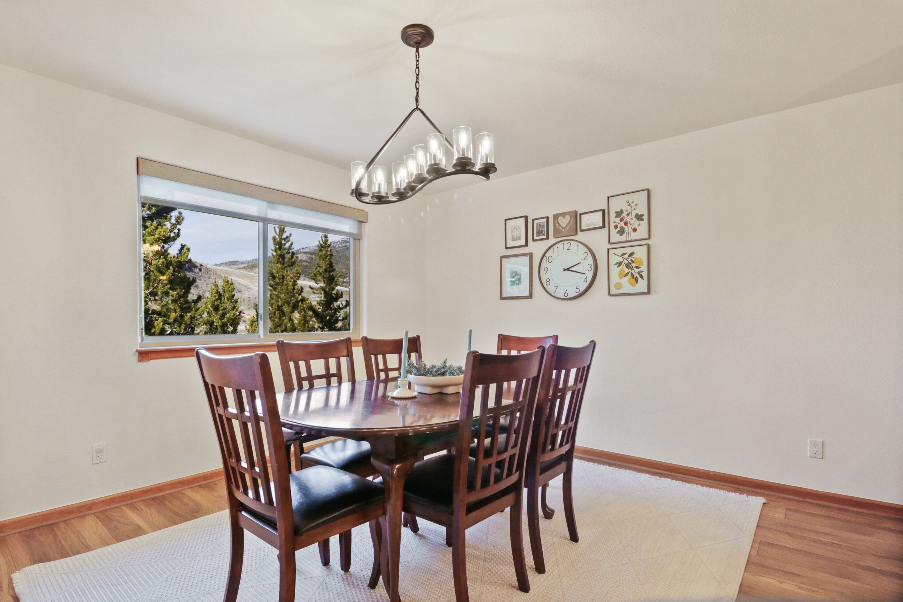 A dining area with a round table, six wooden chairs, a chandelier, a window with a view of trees, and wall-mounted clocks.