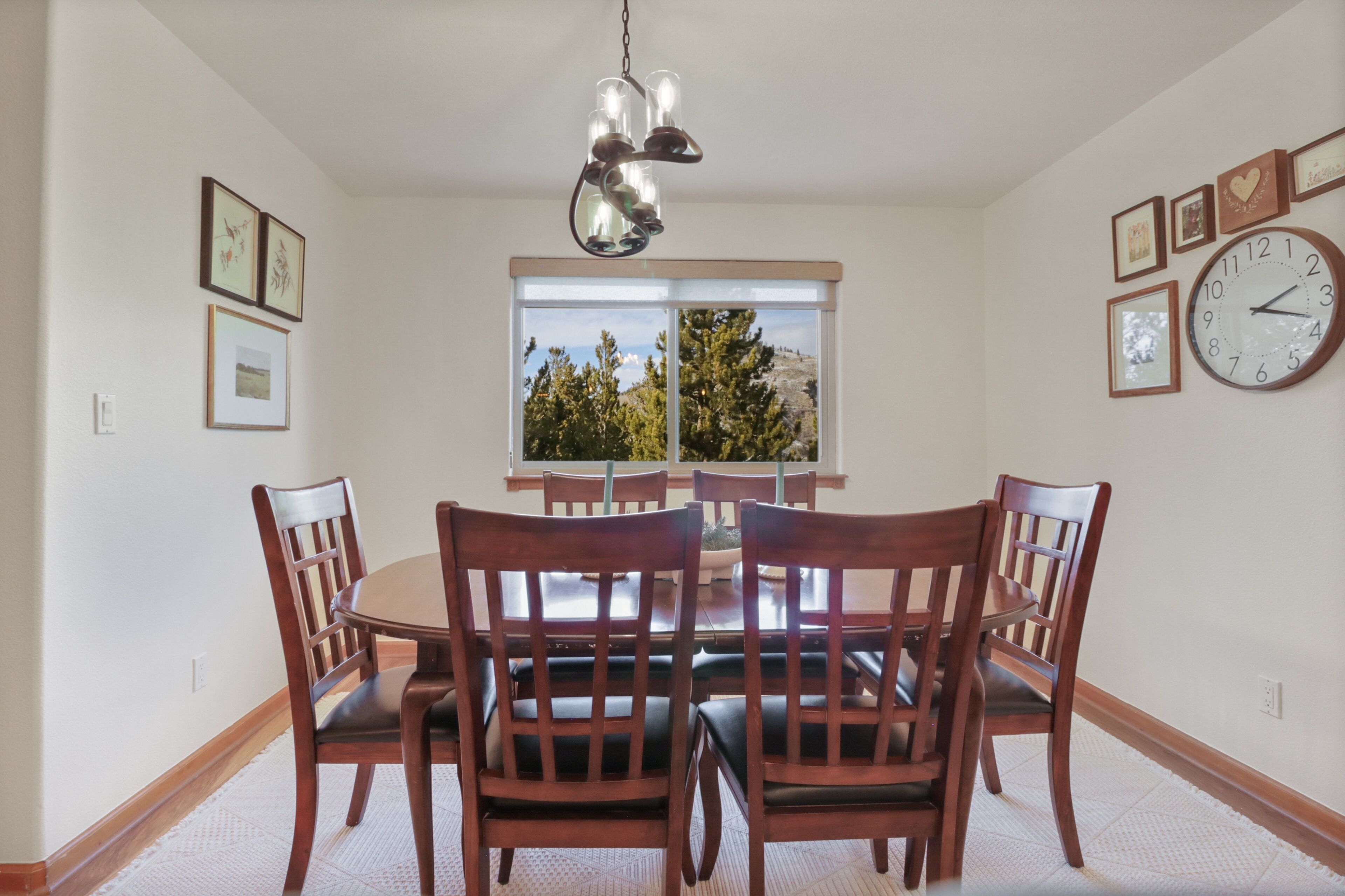 A dining room with a round table and six wooden chairs, a chandelier, and a large window with a view of trees.