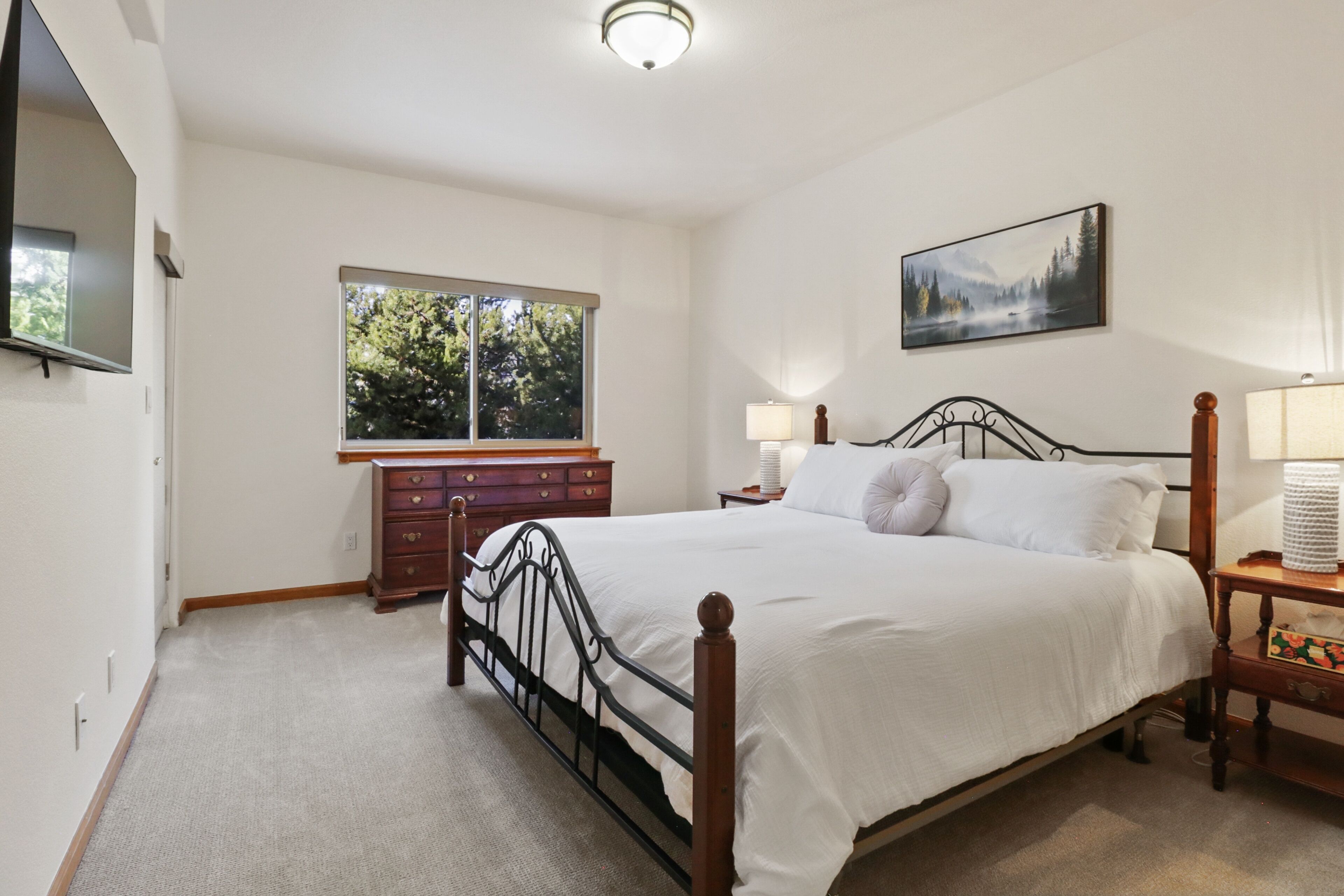 A bedroom with a bed, dresser, nightstand, lamp, and a window with a view of trees.
