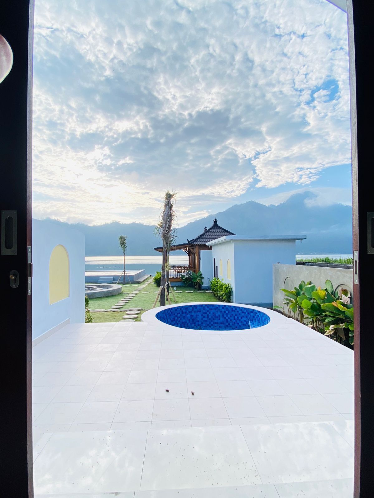A view from a room with a large window overlooking a swimming pool, a small garden, and a distant mountain range.