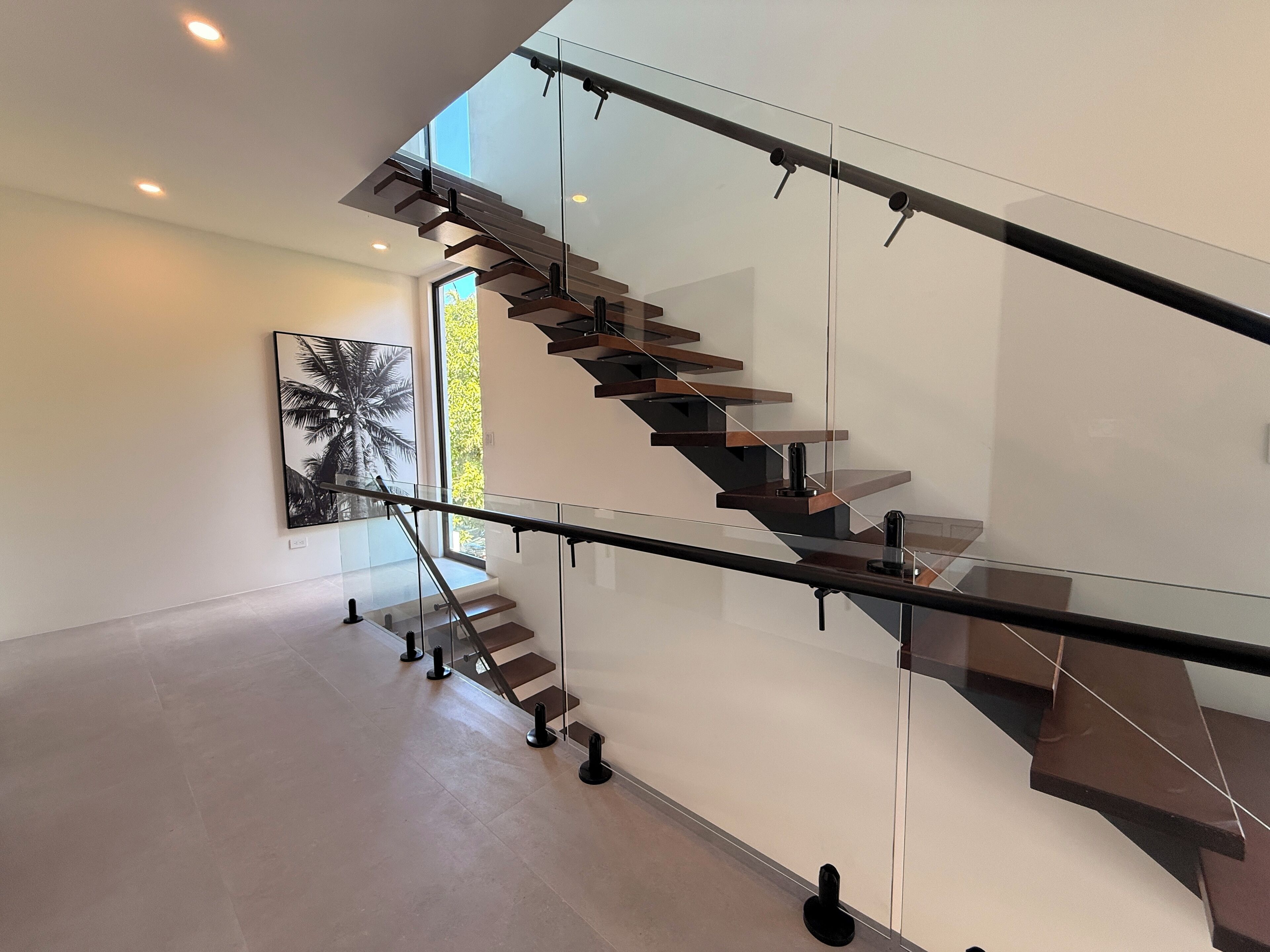 A modern staircase with glass railings and wooden steps, leading to a bright room with a large window and a framed picture on the wall.
