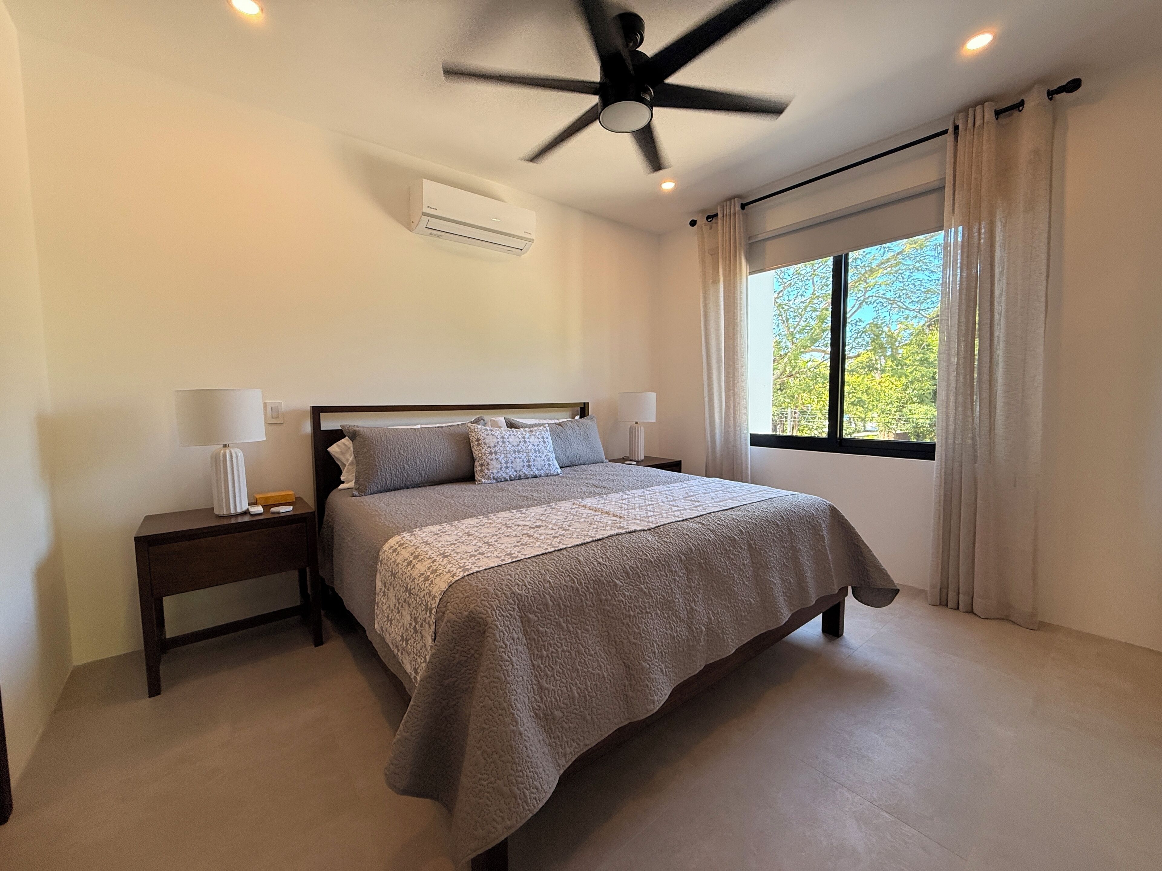 A bedroom with a bed, nightstands, a ceiling fan, an air conditioning unit, and a window with curtains.