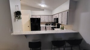 Una cocina moderna con encimera blanca, armarios negros y un refrigerador negro. Hay tres sillas negras en la encimera.