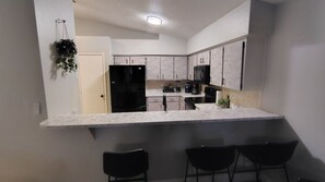 Eine moderne Küche mit weißer Arbeitsplatte, schwarzen Schränken und einem schwarzen Kühlschrank. Drei schwarze Stühle stehen an der Arbeitsplatte.