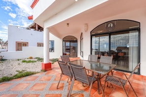 An outdoor patio with a glass table and chairs, a tiled floor, and a view of a building with large windows. - Casa de la Suerte – Ocean View, Las Conchas (Puerto Penasco)