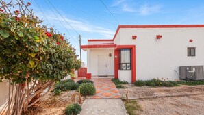 Un edificio con fachada roja y blanca, un camino de hormigón que lleva a la entrada y un pequeño jardín con arbustos y flores.