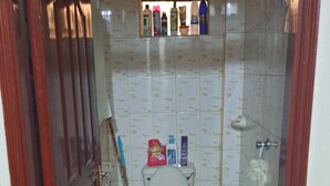 Un baño pequeño con azulejos, que incluye un inodoro, ducha y lavamanos. Se observan productos de limpieza y artículos de aseo en los estantes.