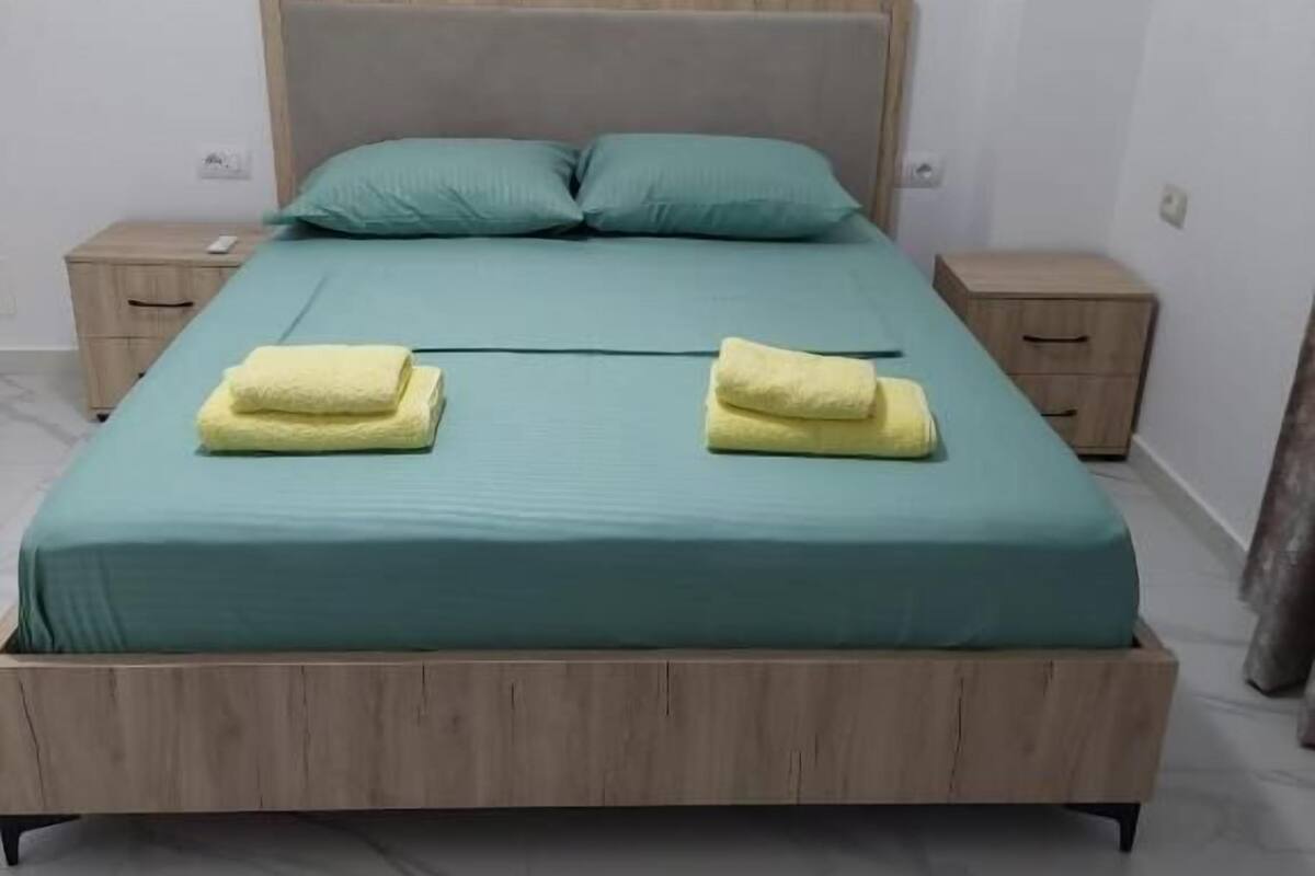 Quarto individual com cama de solteiro, cabeceira de madeira, roupa de cama verde-água e duas toalhas amarelas sobre a cama.