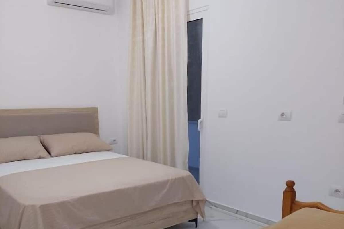 Quarto com cama, mesa de cabeceira, cadeira de madeira, janela com cortinas e um aparelho de ar condicionado na parede.